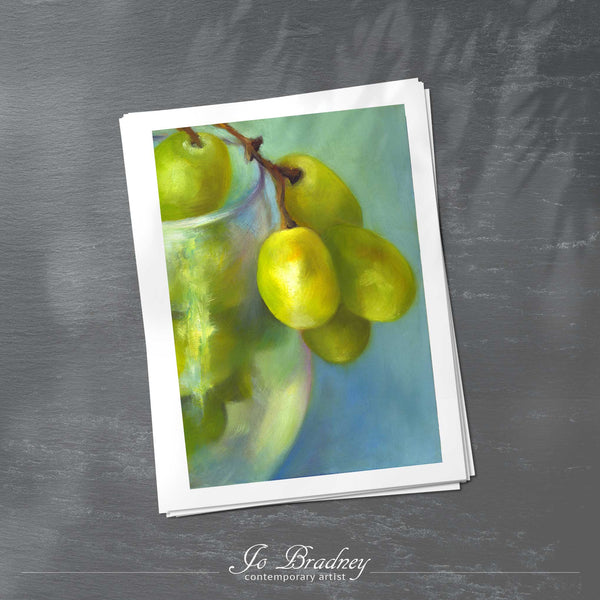 A stack of vertical art prints on archival paper on a slate kitchen counter. The prints show a bunch of green grapes spilling from a white wine glass. This is a giclee print of my realistic oil painting still life. The original artwork is sold.