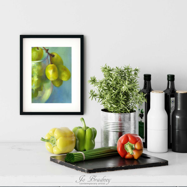 A green grapes oil painting art print in a simple, elegant black picture frame on a kitchen wall. There is a chopping board, vegetables, herbs and some bottles. The art print is as large as three bell peppers. The largest size is 14 inches high by 11 wide