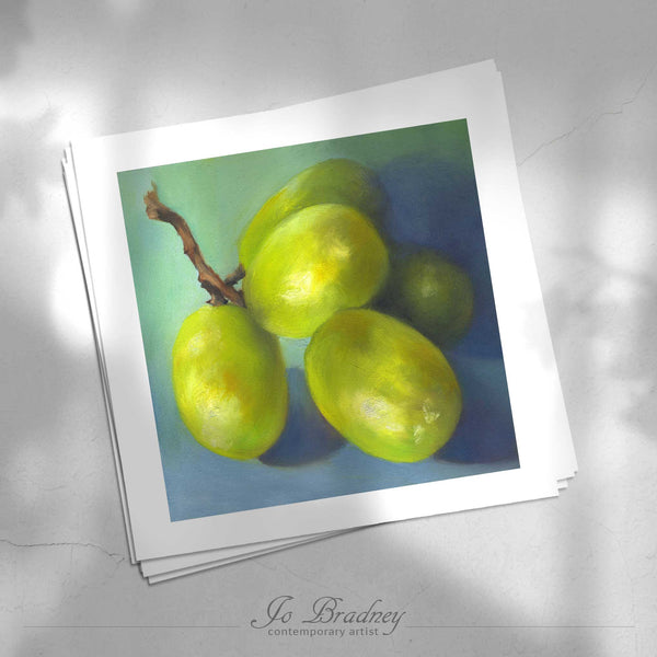 A stack of square art prints on archival paper on a stone kitchen counter. The prints show a bunch of white green grapeson a blue background. This is a giclee print of my realistic oil painting still life. The original artwork is sold.