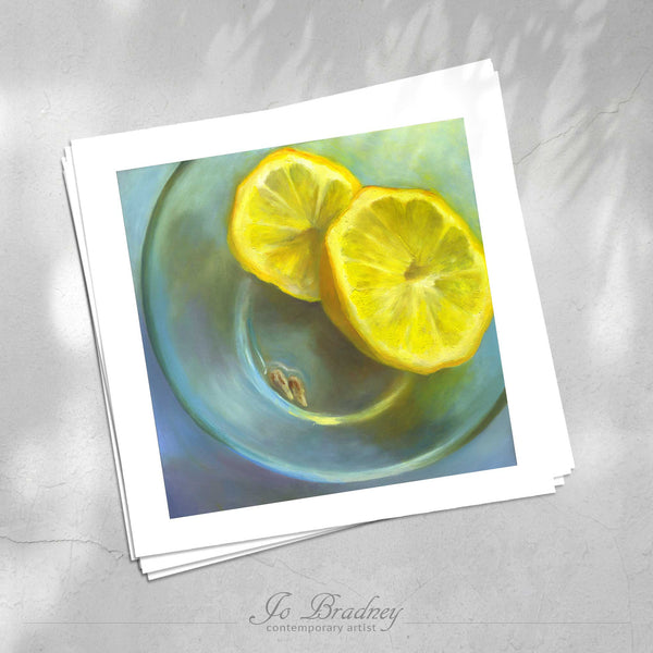 A stack of square art prints on archival paper on a stone kitchen counter. The prints show a yellow lemon and pips in an aqua green glass bowl . This is a giclee print of my realistic oil painting fruit still life. The original artwork is sold.