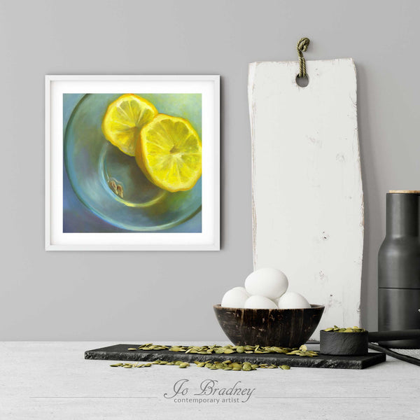 A citrus yellow lemon art print in a simple, elegant white picture frame on a kitchen wall. There is a bowl of eggs, and a chopping board for scale. The print is 5 eggs tall. The smallest square print is 4 inches, the largest is 12 inches.