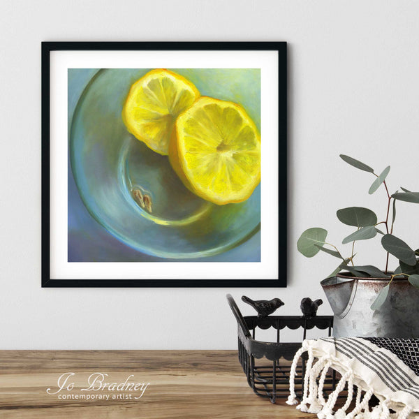 A lemon art print of a fruit oil painting in a simple, elegant black picture frame on a dining room or living room wall. There are flowers in a teapot on a metal tray set on a wood table. The smallest square print is 4 inches, the largest is 12 inches.