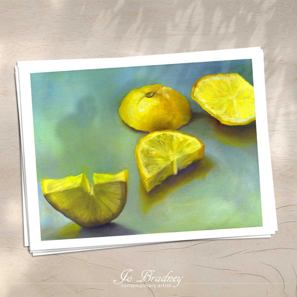 A stack of horizontal art prints on archival paper on a wood kitchen counter. The prints show a fresh cut lemon yellow on a teal green background. This is a giclee print of my realistic oil painting fruit still life. The original artwork is sold.