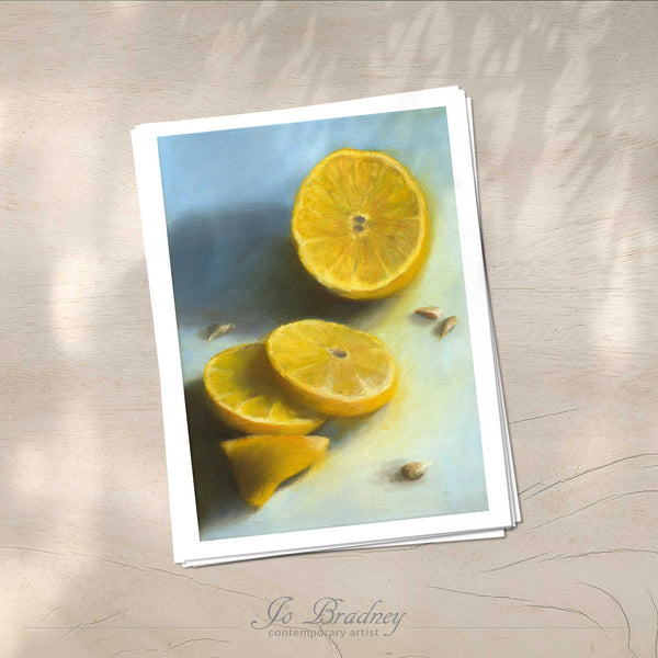 A stack of vertical art prints on archival paper on a wood kitchen counter. The prints show a yellow lemon fresh sliced on an ice blue background. This is a giclee print of my realistic oil painting still life. The original artwork is sold.