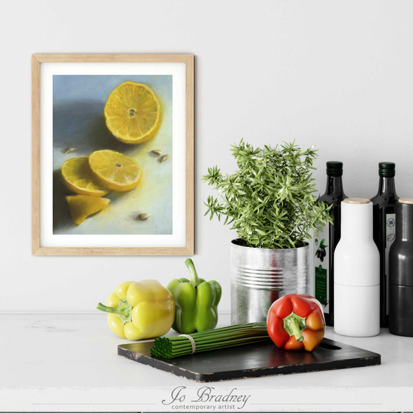 A blueand yellow lemon oil painting print in a simple, elegant wood picture frame on a kitchen wall. There is a chopping board, vegetables, herbs and some bottles. The art print is as large as three bell peppers. The largest size is 14 inches high by 11 wide