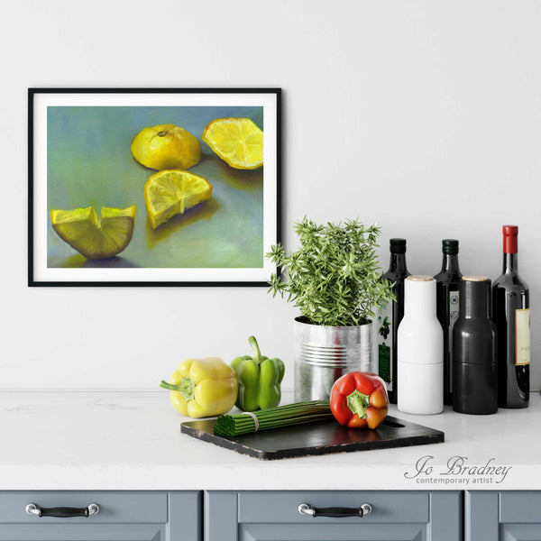A green and yellow lemon painting print in a simple, elegant black picture frame on a kitchen wall. There is a chopping board, vegetables, herbs and some bottles. The art print is as large as three bell peppers. The largest size is 11 inches high by 14 wide