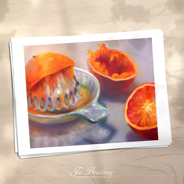 A stack of horizontal art prints on archival paper on a wood kitchen counter. The prints show oranges being squeezed with a glass juicer. This is a giclee print of my realistic oil painting still life.