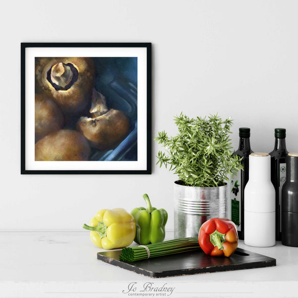 A brown mushroom painting print in a simple, elegant black frame on a modern farmhouse kitchen wall. There is a chopping board, vegetables, herbs and some bottles. The art print is as large as three bell peppers. The largest size is 12 inches square, the smallest is 6 inches.
