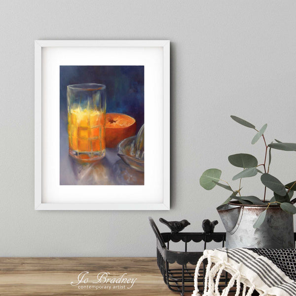 A orange juice art print in a simple, elegant white picture frame on a dining room or breakfast nook wall. There are flowers in a teapot on a metal tray set on a wood table. The smallest vertical print is 4x6 inches, the largest is 11x14 inches.