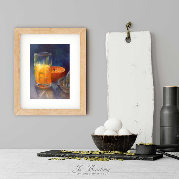 A orange juice art print in a simple, elegant wood picture frame on a modern farmhouse kitchen wall. There is a bowl of eggs, and a chopping board for scale. The print is 5 eggs tall. The smallest vertical print is 4x6 inches, the largest is 11x14 inches.