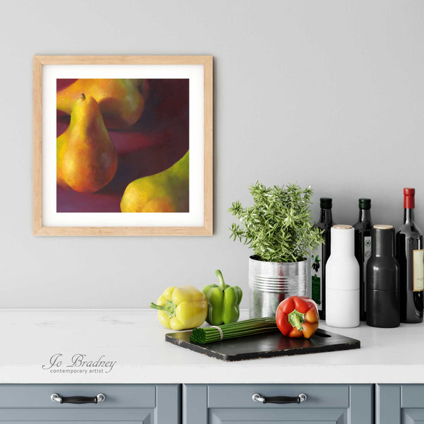 A pear painting giclee art print in a simple, elegant wood picture frame on a kitchen wall. There is a chopping board, vegetables, herbs and some bottles. The art print is as large as three bell peppers. The largest size is 12 inches square, the smallest is4 inches.