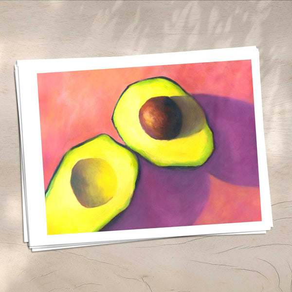 A stack of avocado art prints on archival paper on a wood kitchen counter. The prints show an avocado cut on a bright pink background. This is a giclee print of my realistic oil painting still life. The original artwork is sold.