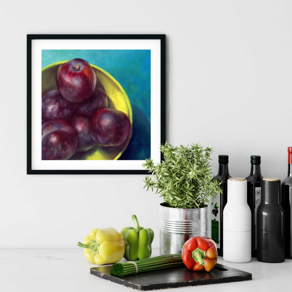 A plum painting giclee art print in a simple, elegant black picture frame on a kitchen wall. There is a chopping board, vegetables, herbs and some bottles. The art print is as large as three bell peppers. The largest size is 12 inches square. the smallest is 4 inches.
