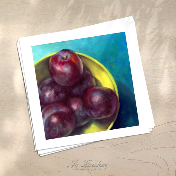 A stack of square art prints on archival paper on a wood kitchen counter. The prints show deep purple plums in a lime green bowl. This is a giclee print of my realistic oil painting still life. 