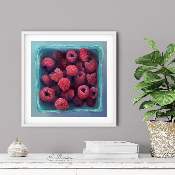 A raspberry oil painting print in a simple elegant white frame on a dining room or living room wall. There are books and a plant in a rustic pot, on a shabby chic painted wood buffet table.