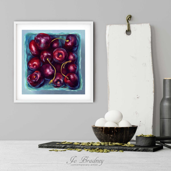 A cherry art print in a simple, elegant white picture frame on a kitchen wall. There is a bowl of eggs, and a chopping board for scale. The print is 5 eggs tall. The smallest squareprint is 4 inches, the largest is 12 inches.