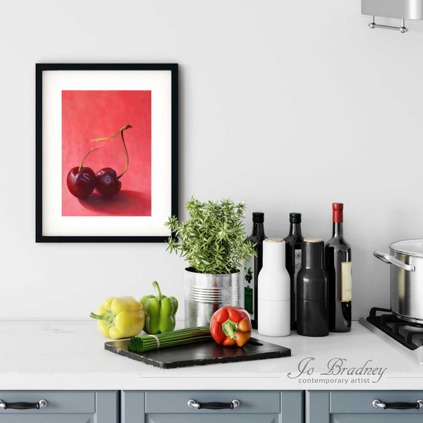 A bright cherry painting art print in a simple, elegant black picture frame on a kitchen wall. There is a chopping board, vegetables, herbs and some bottles. The art print is as large as three bell peppers. The largest size is 14 inches high by 11 wide