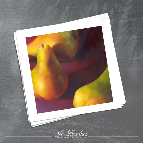 A stack of square art prints on archival paper on a slate kitchen counter. The prints show three yellow pears scattered on a burgundy red background. This is a giclee art print of my realistic oil painting fruit still life. The original artwork is sold.
