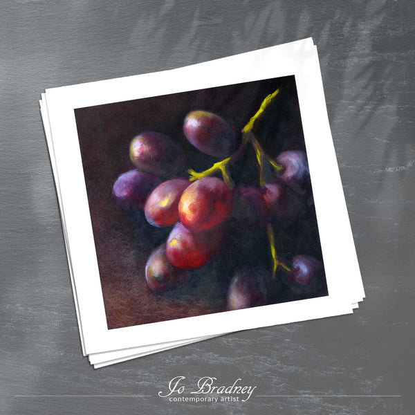 A stack of square art prints on archival paper on a slate kitchen counter. The prints show a bunch of red and purple grapes. This is a giclee print of my realistic oil painting still life. The original artwork is sold.